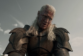Primer plano de un hombre de cabello blanco y largo, con el rostro ensangrentado y cicatrices, vistiendo una armadura oscura contra un cielo nublado