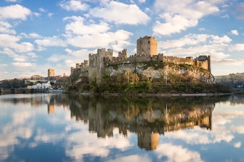 Vista panorámica del Castillo de Pembroke, una fortificación medieval de piedra, reflejada en el agua, bajo un cielo azul con nubes blancas