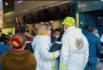Dos personas con chaquetas blancas y detalles reflectantes, un hombre con gorra amarilla y una mujer con carpeta, conversan en un mercado