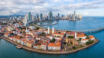 Vista aérea del Casco Viejo de Panamá con tejados rojos y plazas, rodeado por el mar y un viaducto con rascacielos al fondo.