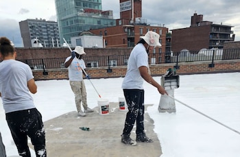 Tres personas utilizan rodillos de pintura con mango largo para cubrir de blanco la superficie de una azotea urbana