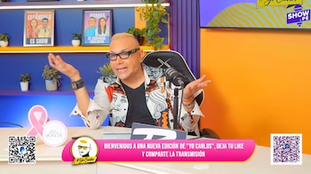 Carlos Cacho sentado ante un micrófono con una camisa estampada y gafas, gesticulando con las manos abiertas en un set azul y naranja