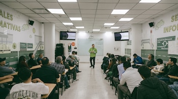 Un instructor de verde se ubica frente a un grupo de personas sentadas en pupitres dentro de un aula decorada con señales de tránsito.
