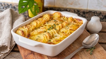 Un recipiente blanco con patatas gratinadas al horno, queso, hierbas verdes. Una cuchara, un salero y un paño en una tabla de madera.