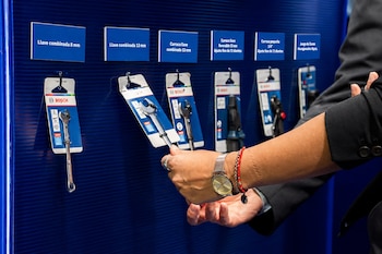 Primer plano de una mano con reloj y pulsera, seleccionando una llave Bosch de un expositor azul. Otras herramientas Bosch y sus etiquetas son visibles