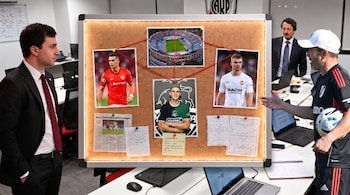 Tres hombres en una oficina con un tablero de corcho que tiene fotos de tres futbolistas, una foto de un estadio, documentos con tácticas y un balón.
