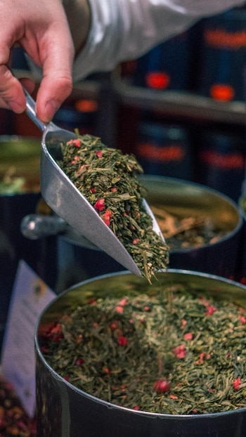 Una mano humana sostiene una pala de metal llena de hojas de té verde con trozos rojos, vertiéndola en un recipiente oscuro y cilíndrico
