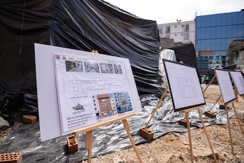 Pizarras con planos arquitectónicos se exhiben en un sitio de construcción con tierra y ladrillos. Al fondo, una lona negra cubre escombros y edificios