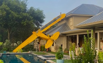 Avión pequeño amarillo incrustado en el tejado de una casa de color claro con un jardín y una piscina azul en primer plano