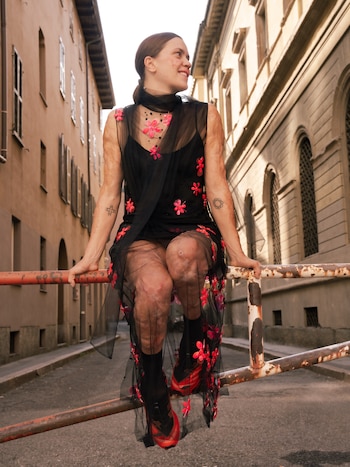 Una mujer vestida con un traje negro con detalles florales rosas se sienta sobre una barandilla metálica en una calle entre edificios