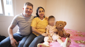 Un hombre, una mujer y una niña con el rostro difuminado sentados en una cama junto a un oso de peluche, con una ventana al fondo