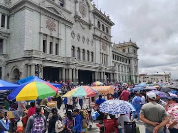 Una multitud de personas con sombrillas de colores se congrega en una calle frente a un edificio grande de piedra con columnas y ventanas arqueadas