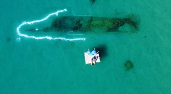 Vista aérea de agua turquesa con forma de submarino delineada con espuma, estructura larga sumergida, balsa flotante con personas y buzos