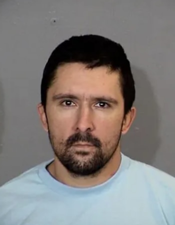 Fotografía policial de un hombre con cabello oscuro, barba corta y camiseta azul claro frente a un fondo gris
