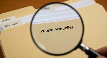 Una mano sostiene una lupa sobre una carpeta de archivo de color beige. A través de la lupa se lee claramente 'Puerto Armuelles'. Otras pestañas dicen 'Puerto Grátas'.