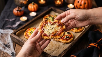 Dos manos sostienen una mini pizza con forma de calabaza frente a una bandeja con más porciones, velas encendidas, calabazas y telarañas.