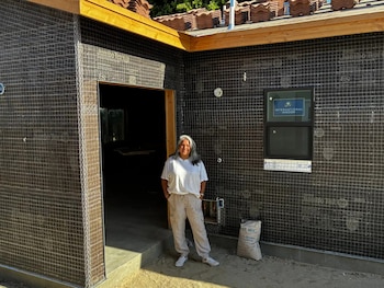 Mujer de pie frente a una pared de casa en construcción cubierta con malla metálica. Hay una ventana instalada y un saco de cemento en el suelo