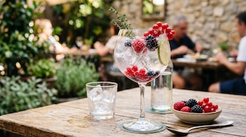 Copa de cristal con bebida con gas, hielo, frutos rojos, lima y una rama de hierba sobre una mesa de madera junto a un cuenco con bayas.