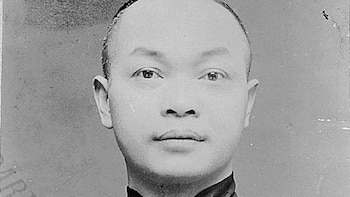 Retrato en blanco y negro de un hombre asiático joven con la cabeza rapada y una vestimenta oscura, mirando directamente a la cámara