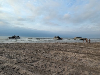 Vista de uma praia arenosa com três navios militares na água e vários soldados na praia sob um céu nublado