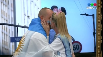 Primer plano de Mariela Prieto y Emanuel Di Gioia besándose, con pintura facial azul y la bandera argentina. Al fondo, otra pareja en pantalla