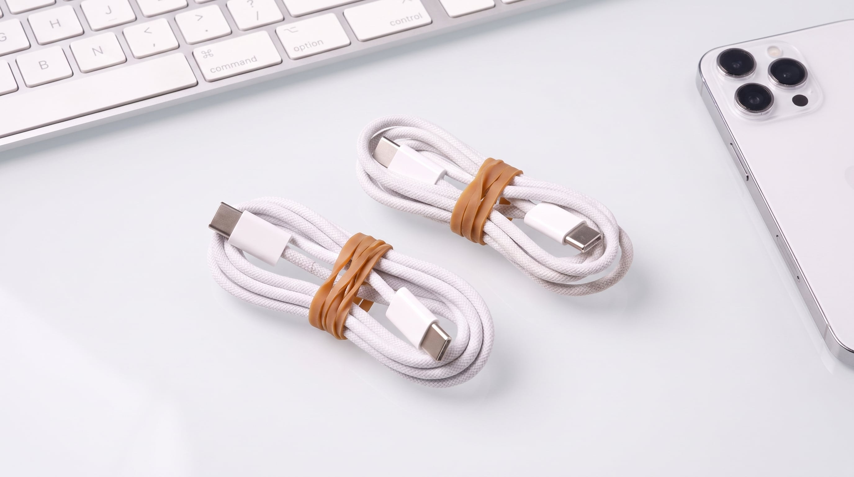Dos cables de carga USB-C enrollados permanecen sujetados por bandas elásticas sobre un escritorio de trabajo con aparatos electrónicos. (Imagen Ilustrativa Infobae)