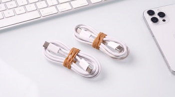 Dos cables blancos con conectores USB-C atados con bandas elásticas junto a un teclado y un teléfono inteligente sobre un escritorio claro.