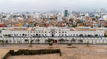 Vista panorámica de un complejo blanco con muros de seguridad junto a una avenida, casas urbanas y cerros bajo un cielo nublado.