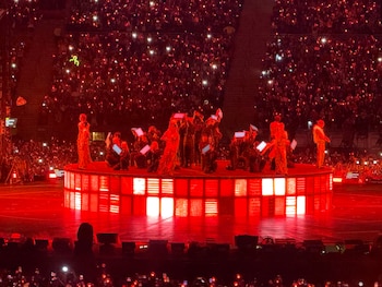 Un escenario circular iluminado de rojo con artistas en el centro, rodeado por tribunas llenas de público con luces rojas encendidas