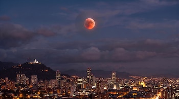 Vista aérea nocturna de ciudad con rascacielos y edificios iluminados, colina con estructura blanca, luna rojiza en el cielo oscuro con nubes.