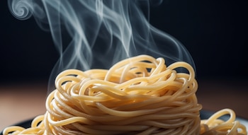 Primer plano de noodles asiáticos cocidos y enrollados, con vapor blanco ascendiendo. El fondo es liso y oscuro, resaltando la comida caliente.