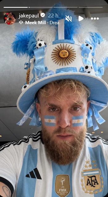 Hombre con barba, camiseta de fútbol celeste y blanca, sombrero con plumas y balones, banderas de Argentina pintadas en las mejillas