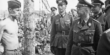 Imagen en blanco y negro: Heinrich Himmler y oficiales nazis cerca de un prisionero sin camisa, demacrado, detrás de una valla de alambre de púas