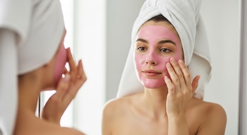 Primer plano de una joven con toalla blanca en la cabeza aplicándose una mascarilla de arcilla rosa en el rostro con los dedos, reflejada en un espejo.