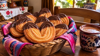 Panes de hojaldre con cobertura de chocolate en una cesta de mimbre junto a una taza decorada con una bebida caliente sobre una mesa.