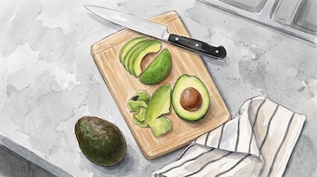 Tabla de madera con aguacate rebanado, la mitad con semilla, cáscaras, un cuchillo, un paño a rayas y un aguacate entero.