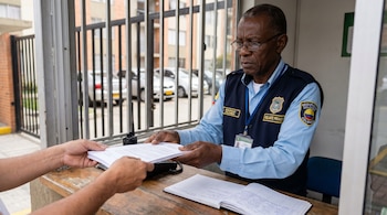 Primer plano de un vigilante de seguridad colombiano de piel negra con uniforme y gafas, recibiendo documentos de otra persona en una portería con reja y registro.