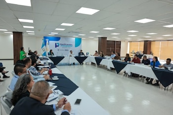 Sala de reuniones con personas sentadas alrededor de una mesa en U, con computadoras, documentos y tazas. Un banner con "Planificación y Presupuesto Abierto 2021" y banderas al fondo