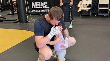 Un hombre con las manos vendadas se agacha junto a una niña pequeña y le cubre el rostro con la mano en un gimnasio.