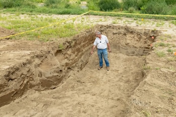 Un hombre con camisa estampada y jeans está de pie en una excavación profunda, señalando una pared de tierra. El área superior tiene cinta amarilla