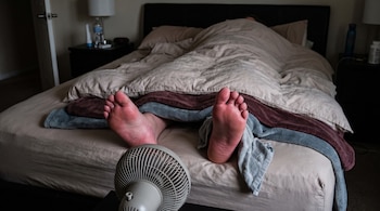Dos pies de persona asoman de la ropa de cama sobre una cama. Un ventilador pequeño dirige aire hacia los pies. A un lado, una toalla gris.