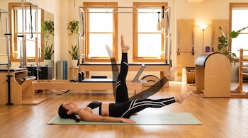 Mujer con ropa deportiva haciendo pilates en un tapete verde, sus piernas en movimiento, en un estudio con máquinas de pilates, ventanales y plantas.