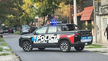 Una camioneta negra de la Policía de Santa Fe con luces azules encendidas bloquea una calle residencial arbolada en un día nublado; un oficial camina por la vereda junto a edificios