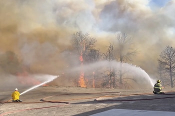 Dos bomberos arrodillados rocían agua sobre un gran incendio forestal con humo denso y árboles en llamas en el fondo