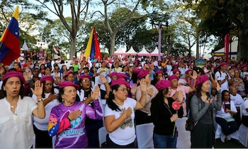 Varias mujeres con boinas rosadas levantan una mano o la llevan al pecho en un evento al aire libre con banderas de Venezuela al fondo