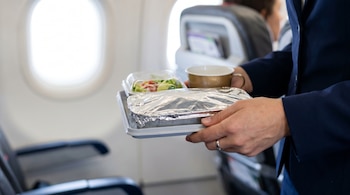 Primer plano de manos femeninas sosteniendo una bandeja de comida de avión con ensalada, plato principal y una taza, dentro de la cabina de un avión.