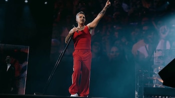 Robbie Williams canta en un escenario con ropa roja, sostiene el parante de un micrófono y apunta hacia arriba con la mano