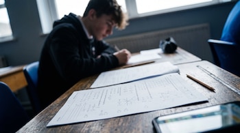 Examen impreso con gráficos sobre una mesa de madera, un lápiz, un teléfono celular y un estudiante fuera de foco al fondo.
