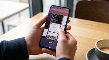 Dos manos sostienen un teléfono inteligente Android que muestra varias aplicaciones abiertas en segundo plano, incluyendo Spotify, Google Chrome e Instagram. Una taza de café está a la derecha.