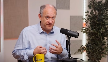 Adam Fox, un hombre de mediana edad con camisa azul claro, habla ante un micrófono negro. Una taza amarilla y una planta verde son visibles